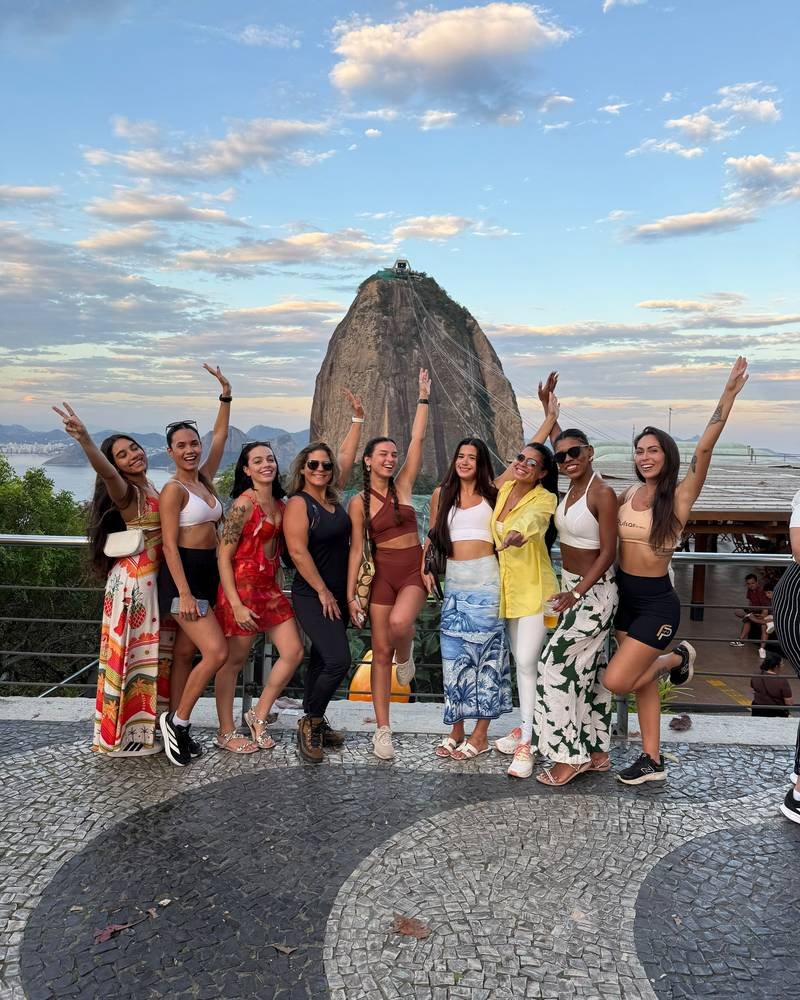 Nove participantes de braços erguidos em um mirante, com o Pão de Açúcar e o céu do fim de tarde ao fundo.