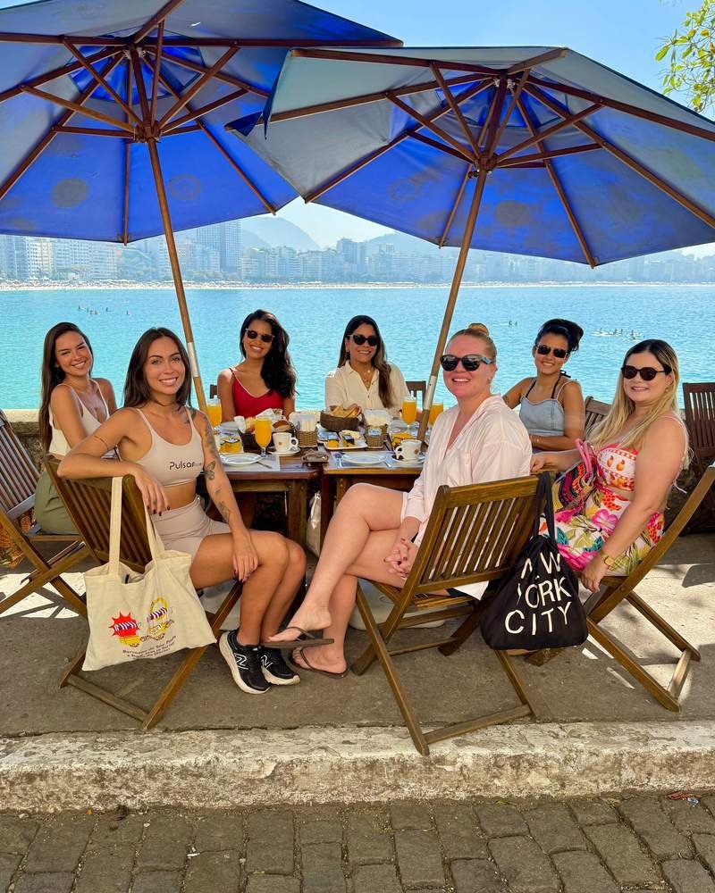 Sete participantes sentadas em mesas sob guarda-sóis azuis, tomando café da manhã com a orla e o mar logo atrás.