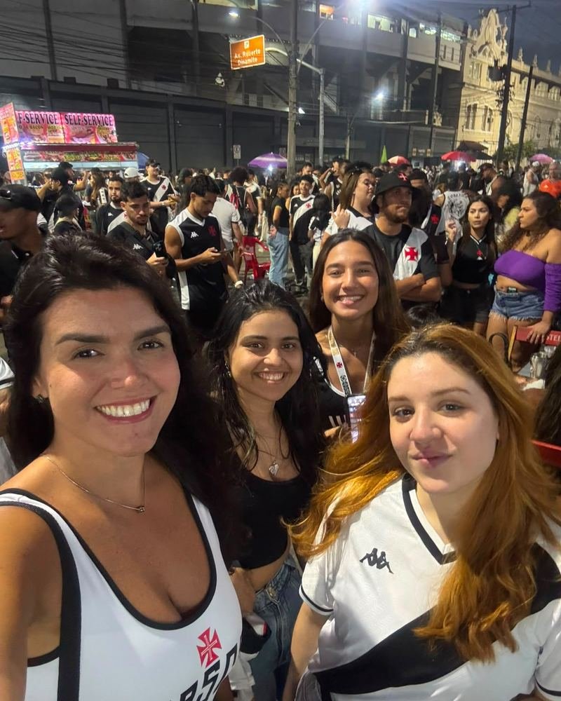 Quatro participantes com camisa do Vasco tirando uma selfie na rua, com a torcida e o estádio ao fundo antes do jogo.